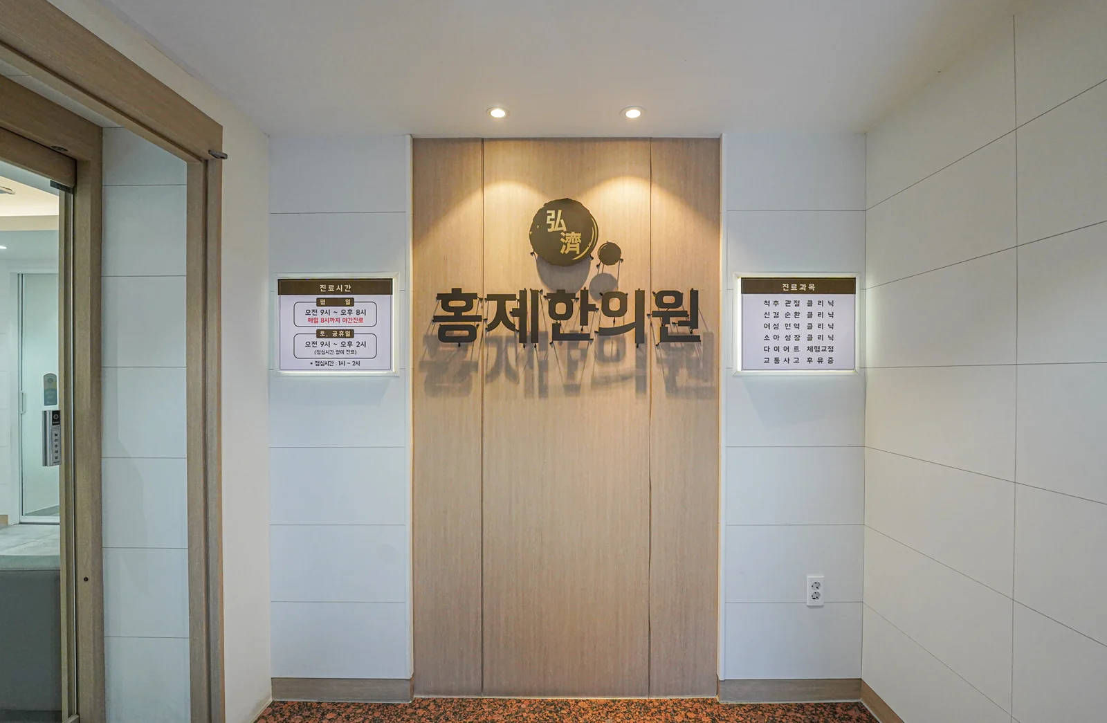 나무 벽에 「弘濟 홍제한의원」 사인을 붙인 전실, 좌우에 진료시간·진료과목 안내판이 걸려 있다